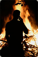 Picture of man standing in the dark outside of his phone. There is a fire behind him