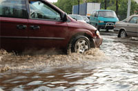 Picture of a van sitting in flooded water on the road. There is other vehicles going through water on the road in the background